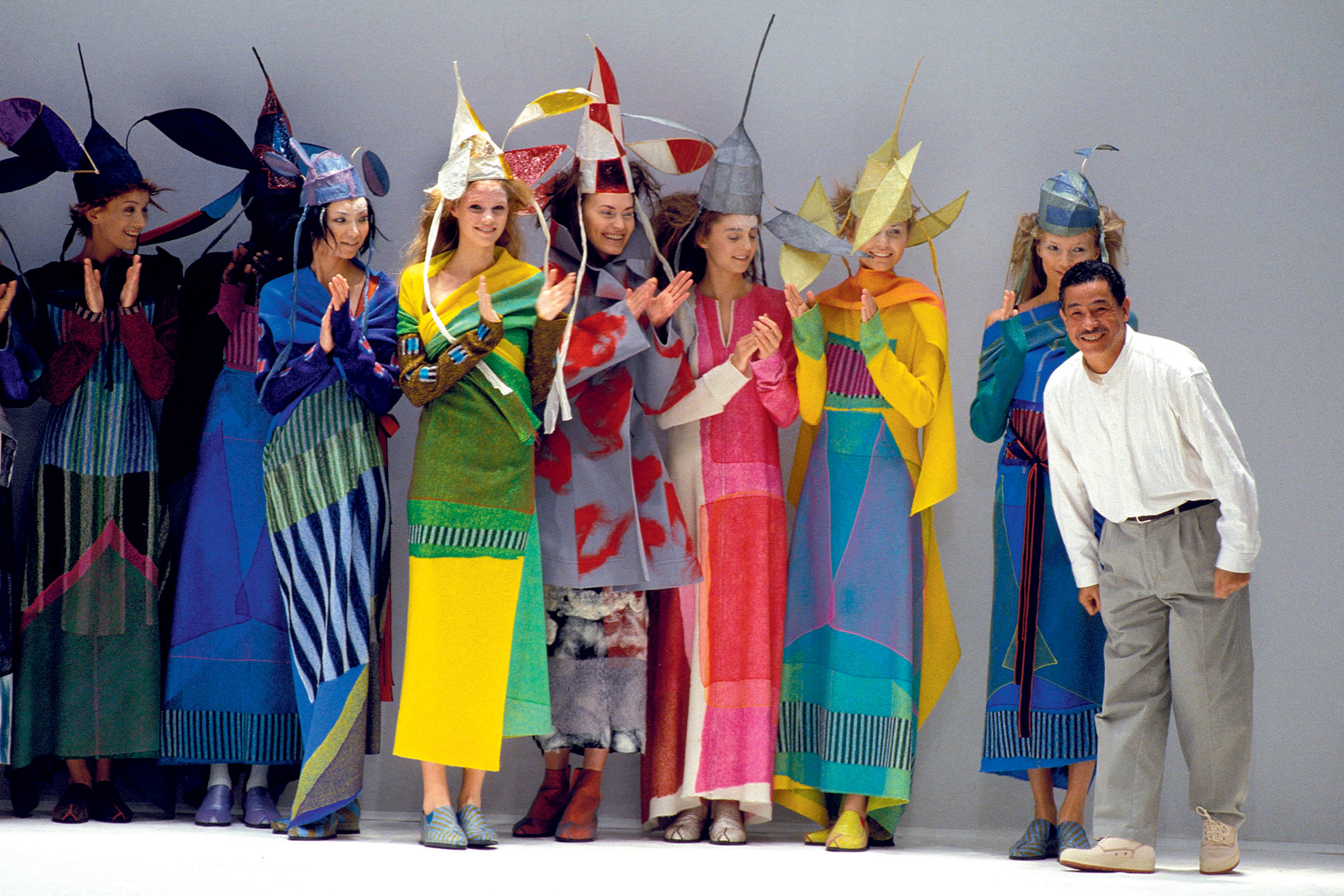 O estilista Issey Miyake, sorrindo e curvado, ao lado de oito modelos aplaudindo, vestindo roupas coloridas e chap&eacute;us esculturais em um desfile de moda