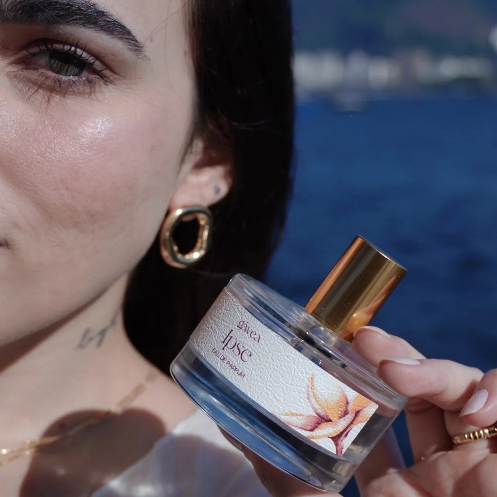 Close-up de uma mulher com cabelo escuro e brinco dourado, segurando um frasco de perfume G&aacute;vea Ipse Eau de Parfum com r&oacute;tulo floral, contra um fundo azul desfocado