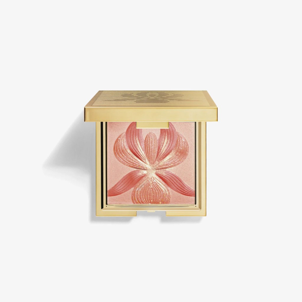 Estojo quadrado dourado de maquiagem aberto, revelando um iluminador em p&oacute; rosa p&ecirc;ssego com relevo de flor de l&oacute;tus e brilho dourado