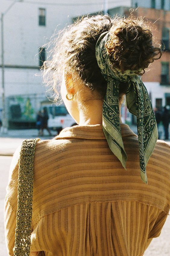 Pessoa de costas, cabelo cacheado preso com len&ccedil;o verde estampado, camisa listrada marrom e brinco dourado, em rua urbana