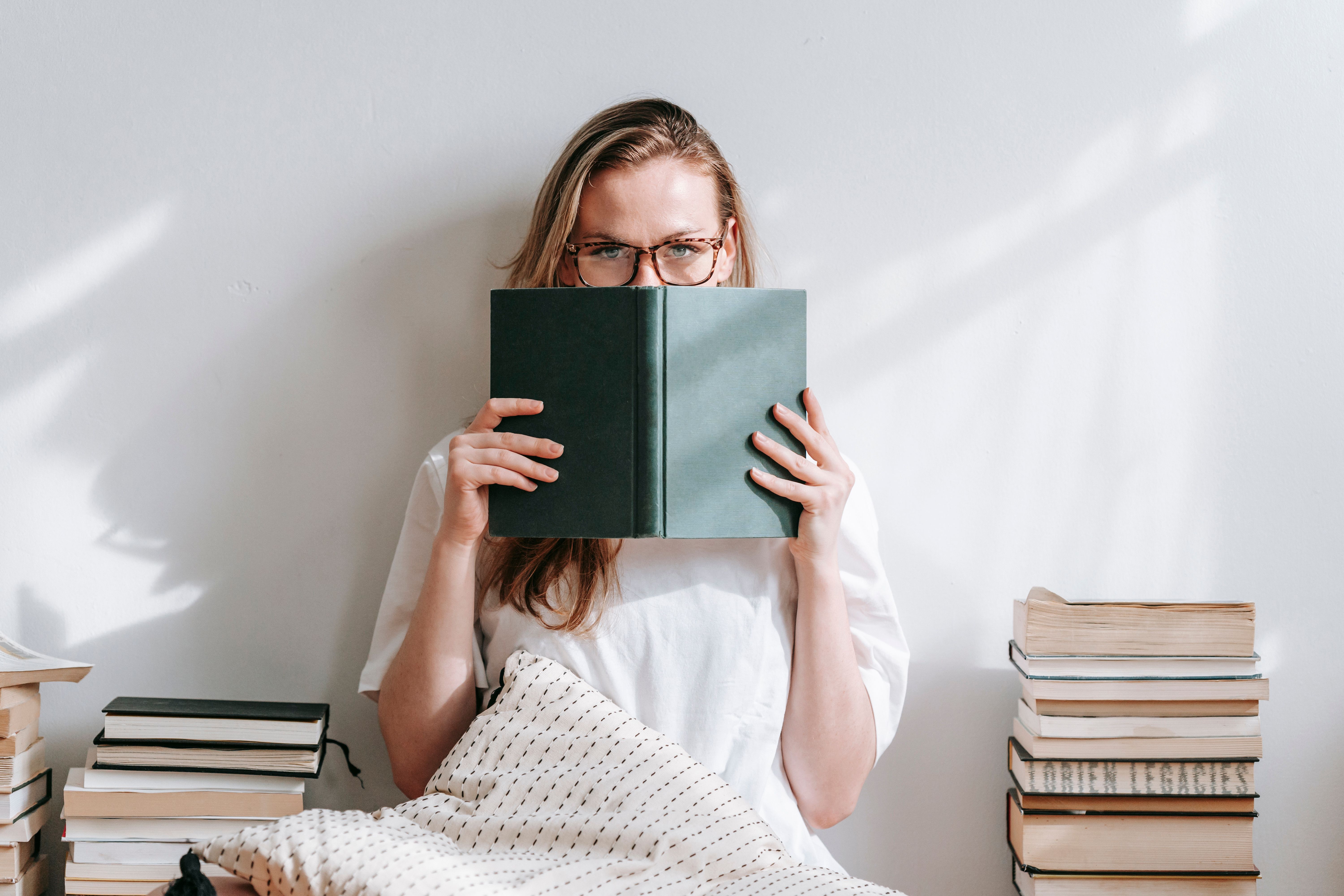 Mulher de óculos e camiseta branca esconde o rosto atrás de um livro verde escuro, sentada no chão com almofada e pilhas de livros ao redor