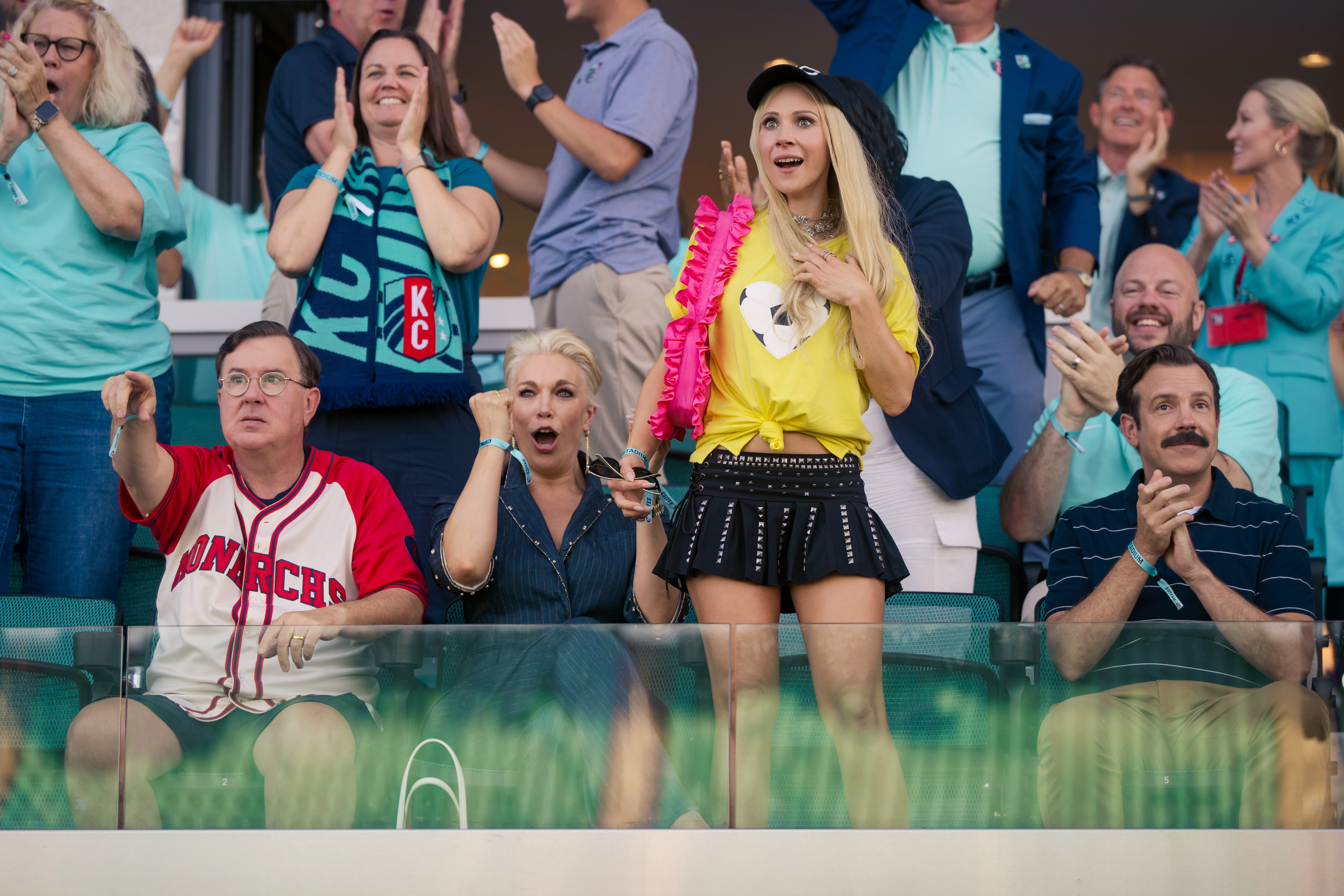 Pessoas assistem a um jogo em um est&aacute;dio. Um homem de &oacute;culos e camisa de beisebol branca e vermelha aponta para frente. Ao lado, uma mulher loira de boca aberta e uma jovem loira de blusa amarela e minissaia preta olham para cima. Outros torcedores aplaudem ao fundo, incluindo um homem de bigode e camisa polo listrada