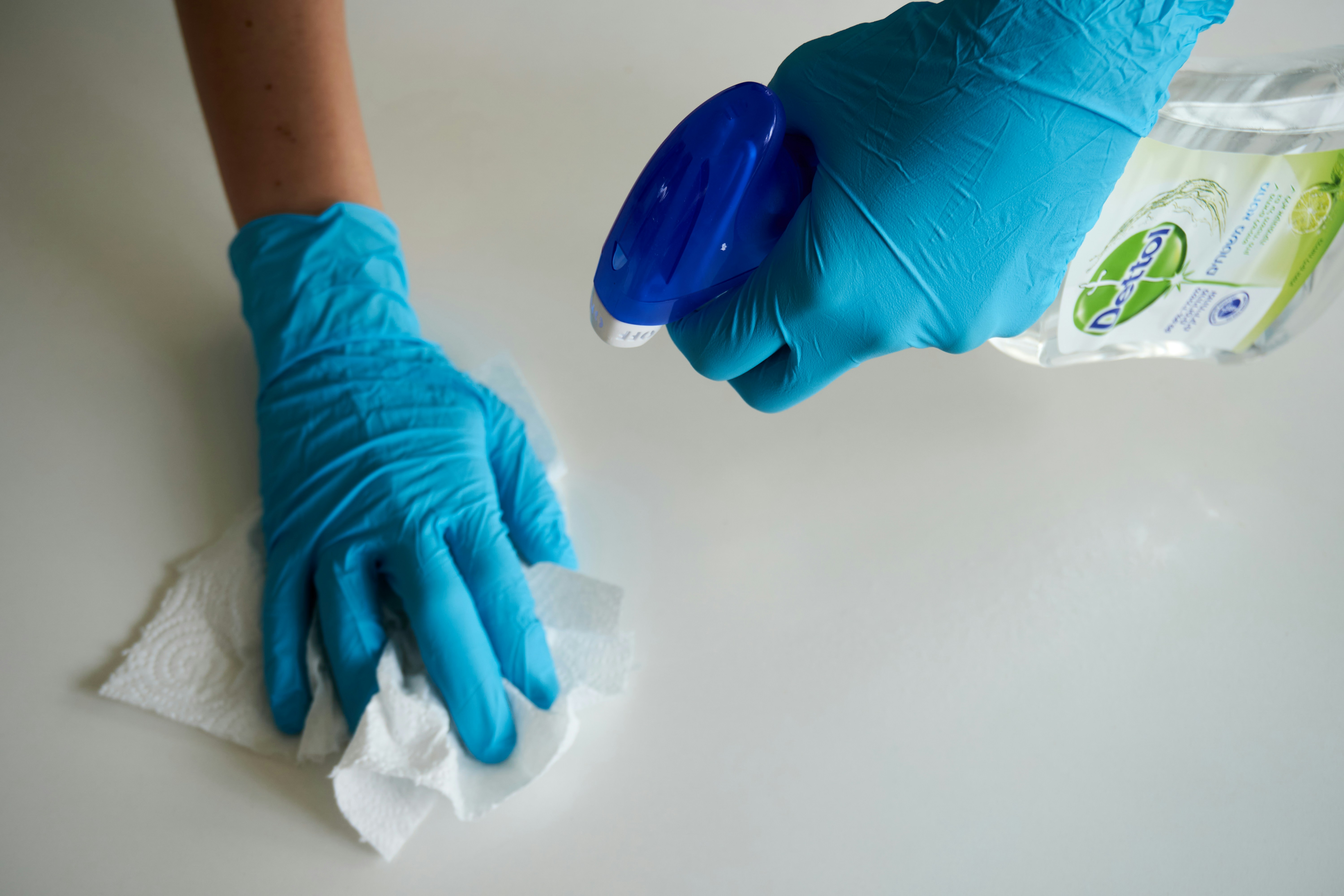 Duas m&atilde;os com luvas azuis, uma segurando um borrifador azul e transparente com l&iacute;quido Dettol, e a outra limpando uma superf&iacute;cie branca com papel toalha