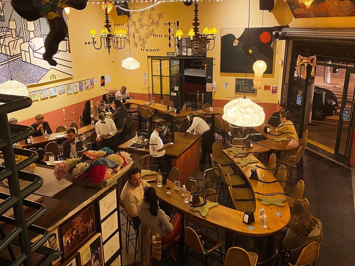 Vista a&eacute;rea de um restaurante com mesas de madeira e cadeiras de palha, iluminado por luzes quentes e pendentes. Clientes conversam em grupos e casais, enquanto gar&ccedil;ons servem. Uma parede tem um mural de arte pop e outra, um quadro abstrato. Ao fundo, a rua noturna com um carro estacionado