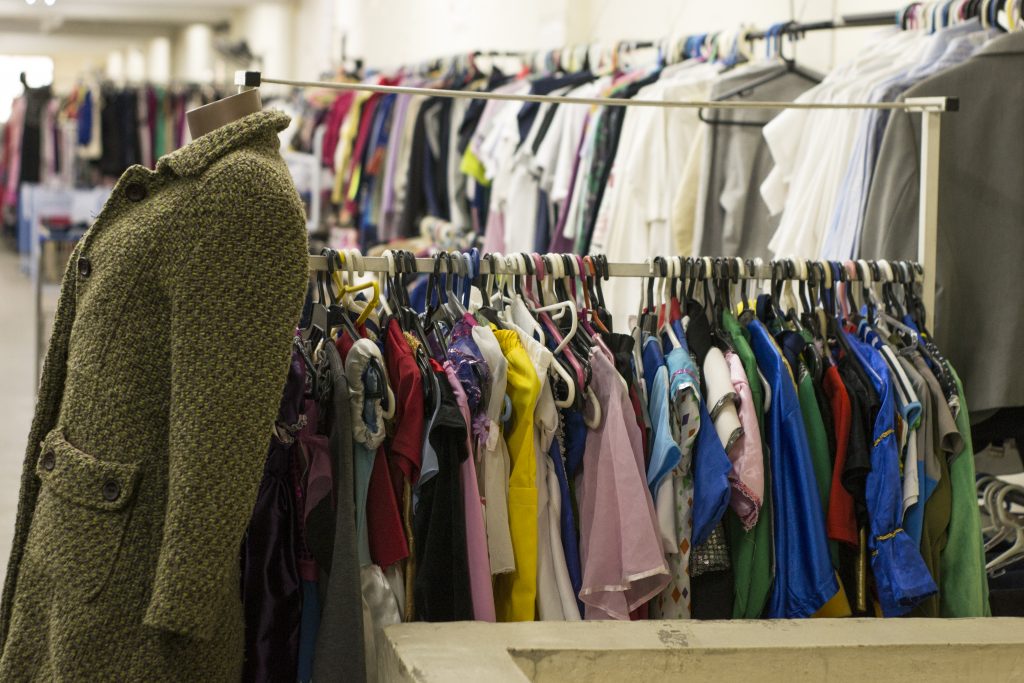 Mannequim com casaco de tweed verde-oliva em primeiro plano, &agrave; esquerda, e araras cheias de roupas coloridas e variadas ao fundo, em um brech&oacute; ou loja de departamento