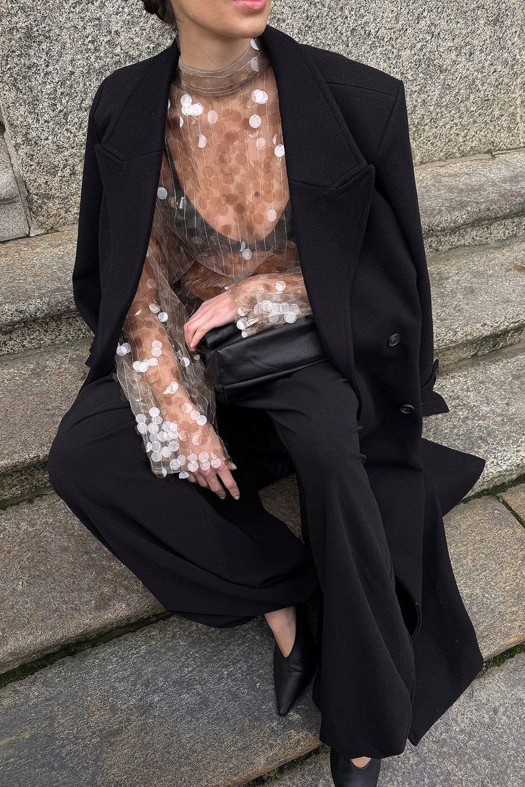 Mulher sentada em escada de pedra, vestindo casaco preto, blusa transparente com bolinhas brancas, cal&ccedil;a preta e sapatos pretos. Ela segura uma bolsa preta no colo