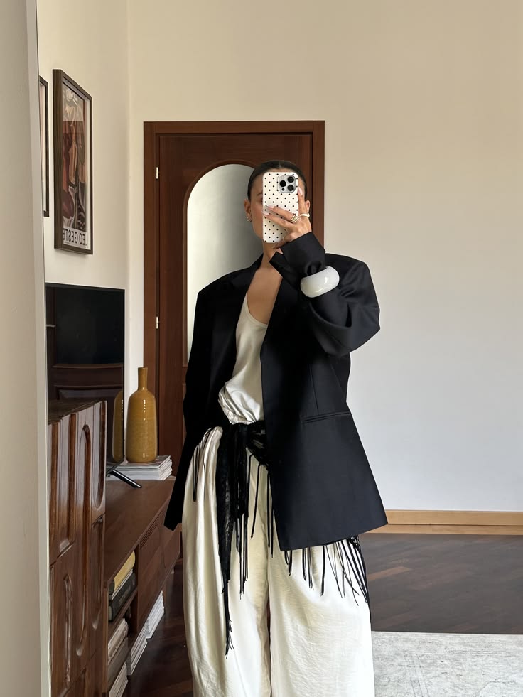 Pessoa de p&eacute; em frente a um espelho, tirando uma selfie com o rosto coberto pelo celular. Veste blazer preto oversized, blusa branca de seda, cal&ccedil;a pantalona branca e cinto preto com franjas. Usa pulseira branca e an&eacute;is dourados. Ao fundo, um m&oacute;vel de madeira e uma porta marrom com arco
