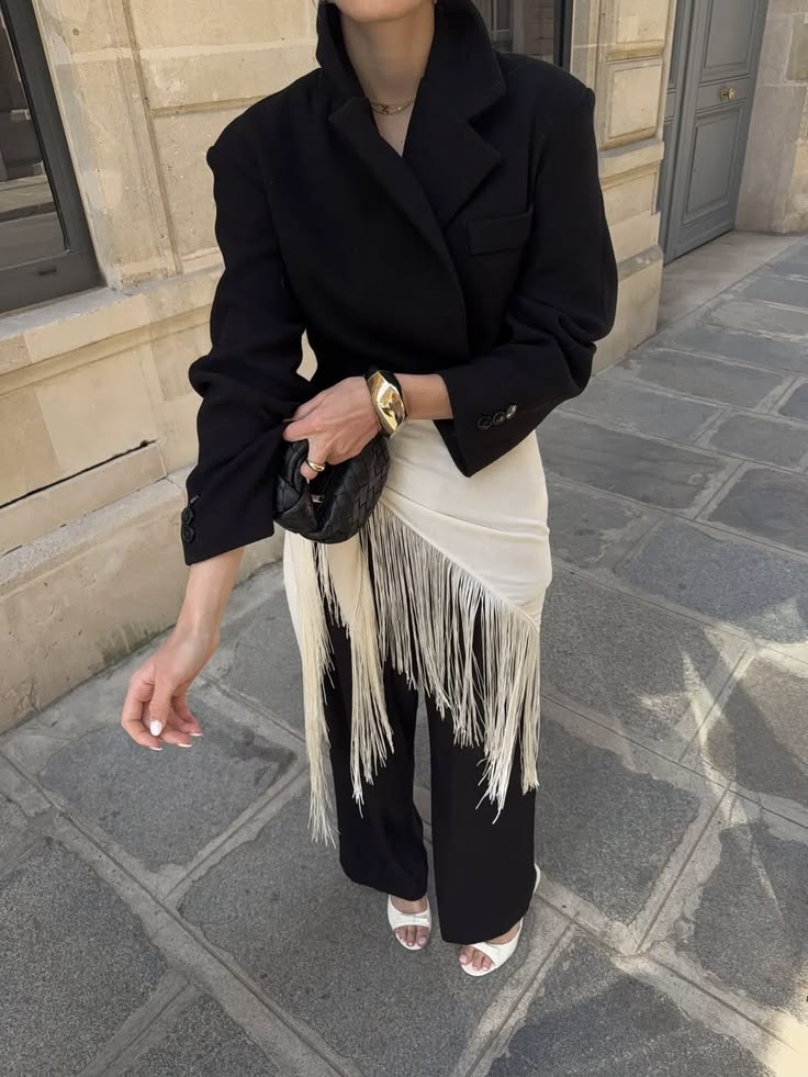 Pessoa de p&eacute;, vestindo blazer preto, cal&ccedil;a preta, sand&aacute;lias brancas e uma saia creme com franjas sobre a cal&ccedil;a. Segura uma bolsa preta pequena e usa um bracelete dourado no pulso esquerdo, em um piso de pedras com um pr&eacute;dio ao fundo