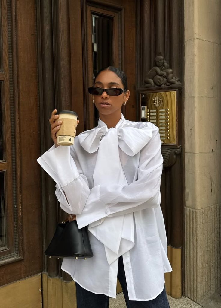 Mulher de pele morena, &oacute;culos escuros e camisa branca com la&ccedil;o grande no pesco&ccedil;o, segurando um copo de caf&eacute; e uma bolsa preta, em frente a uma porta de madeira e interfone dourado