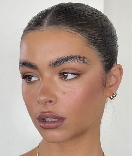 Rosto de mulher jovem com pele bronzeada, cabelo escuro preso, maquiagem natural com blush rosado e batom nude, olhando para a direita. Ela usa um brinco dourado com pedras brancas e um colar fino.