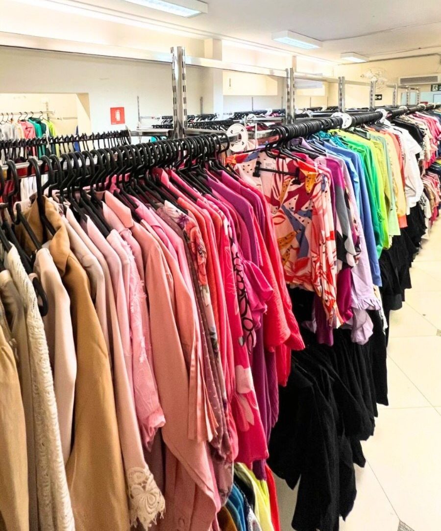 Corredor de loja com araras cheias de roupas coloridas, organizadas por tons. &Agrave; esquerda, pe&ccedil;as em bege e rosa claro; ao centro, tons de rosa, roxo e estampas florais; &agrave; direita, roupas em verde, azul e preto. O teto branco tem lumin&aacute;rias retangulares e trilhos met&aacute;licos.