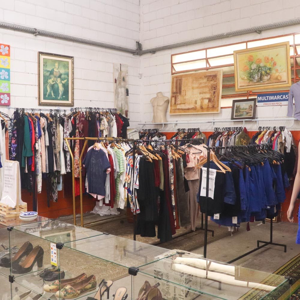 Interior de um brech&oacute; com ar r&uacute;stico, paredes brancas de tijolos e tubula&ccedil;&otilde;es expostas. V&aacute;rias araras de roupas coloridas e variadas, como blusas, cal&ccedil;as e vestidos, preenchem o espa&ccedil;o. Quadros decorativos e um busto masculino adornam as paredes. No primeiro plano, um balc&atilde;o de vidro exibe sapatos femininos e outros itens. Um tapete estampado cobre parte do ch&atilde;o, e a ilumina&ccedil;&atilde;o &eacute; clara