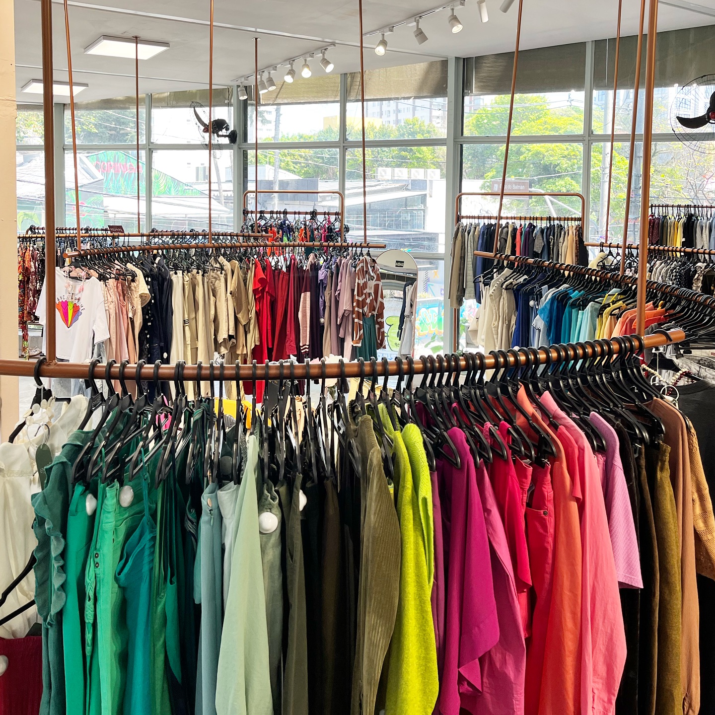 Araras de roupas coloridas em uma loja, com pe&ccedil;as organizadas por tons vibrantes de verde, azul, rosa e vermelho, e outras araras ao fundo com roupas em tons neutros e variados. Grandes janelas ao fundo revelam &aacute;rvores e pr&eacute;dios externos