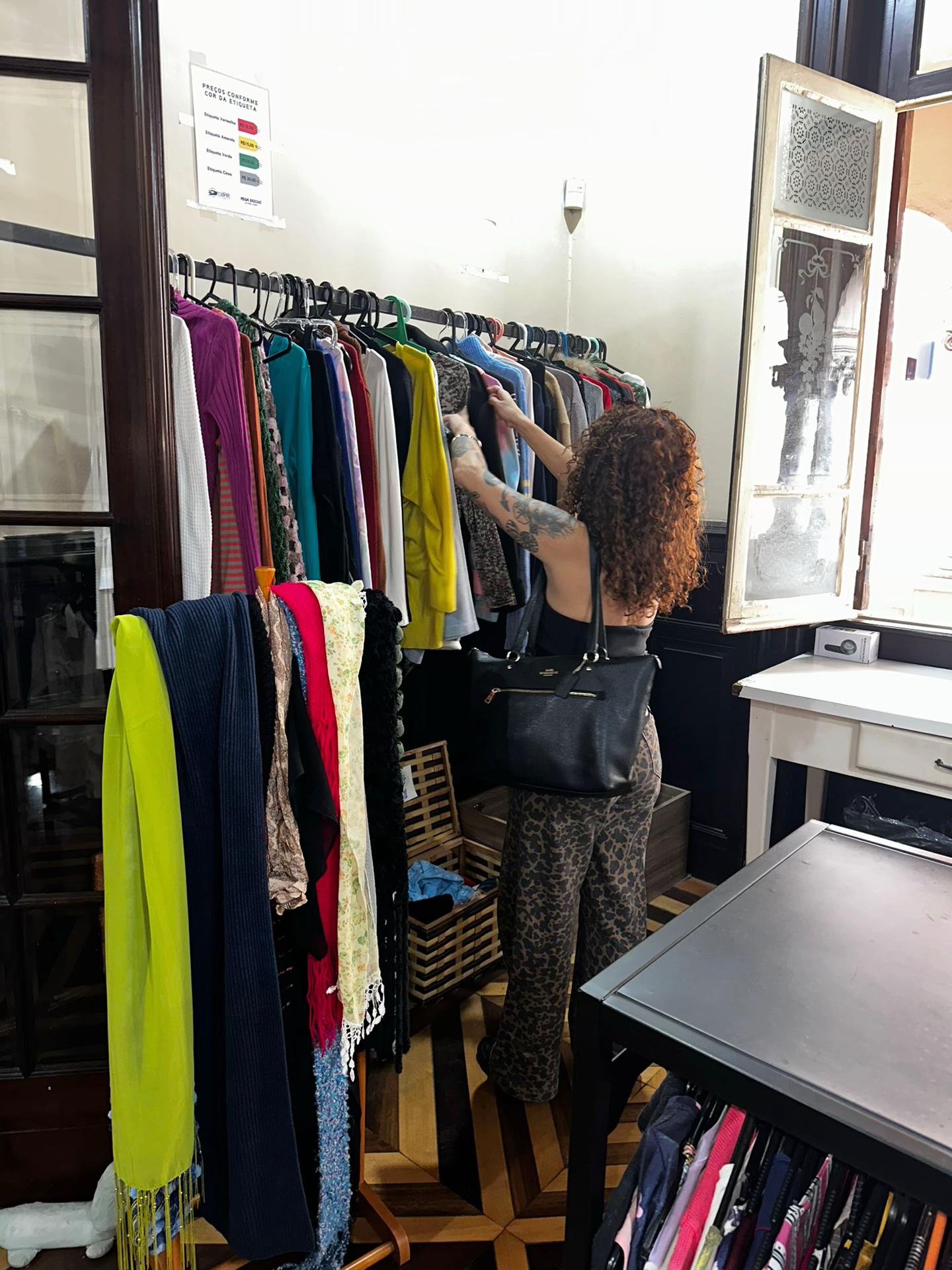 Mulher de cabelos ruivos cacheados, blusa preta e cal&ccedil;a estampada de oncinha, olhando roupas em um cabideiro de brech&oacute;. Ela segura uma bolsa preta no ombro e tem tatuagens no bra&ccedil;o esquerdo. Ao fundo, uma janela antiga e um aviso de pre&ccedil;os por etiqueta.