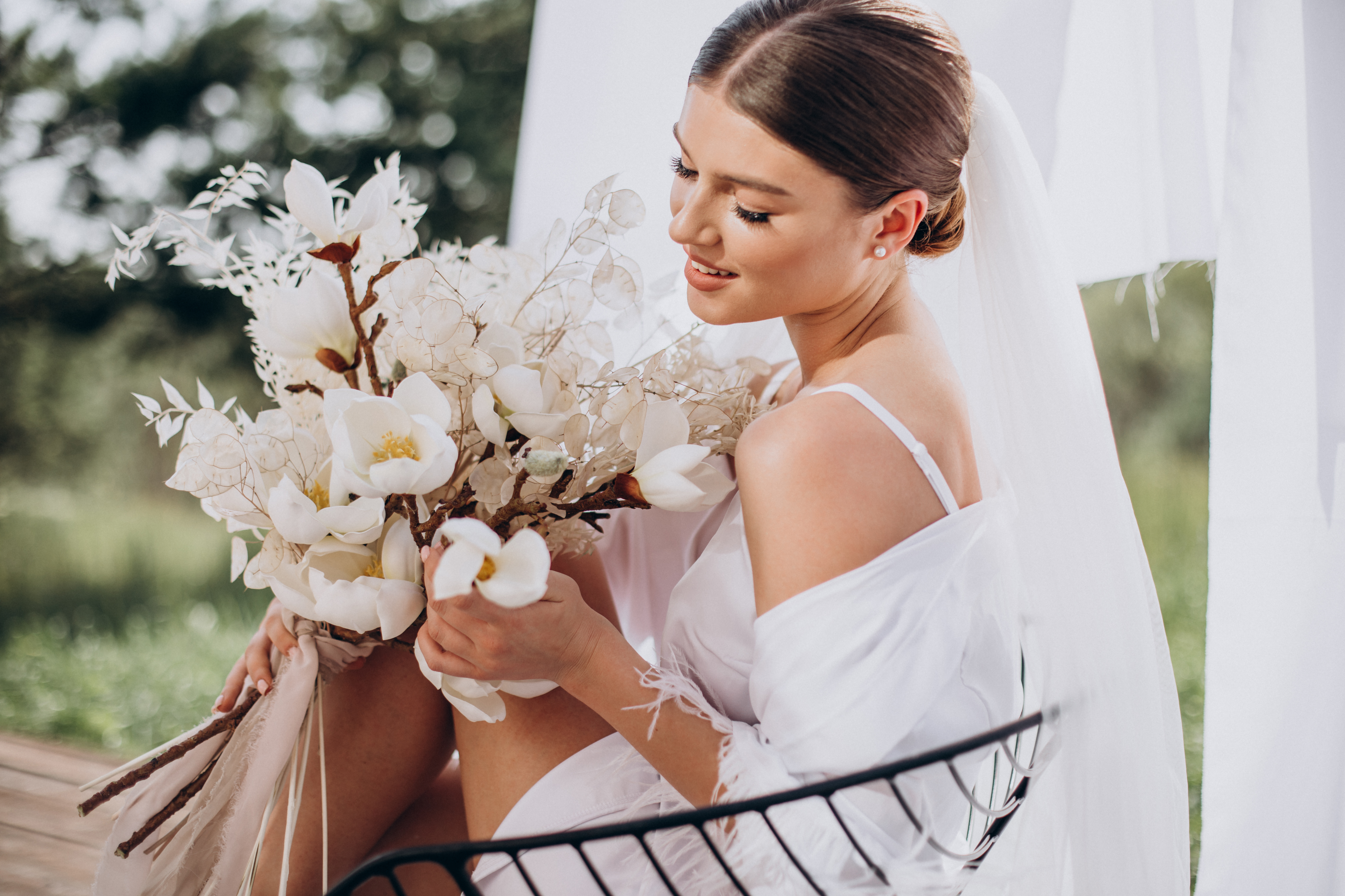Noiva jovem, com v&eacute;u e coque, sorri enquanto segura um buqu&ecirc; de flores brancas e folhagens secas, vestindo um robe branco sobre uma al&ccedil;a fina, sentada em uma cadeira de ferro com cortinas claras ao fundo