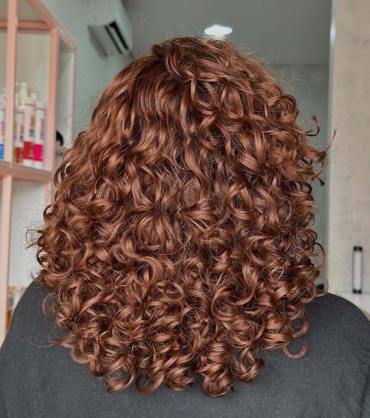 Cabelo cacheado ruivo vibrante, com cachos definidos e volumosos, visto de costas, sobre ombros de uma pessoa vestindo blusa preta