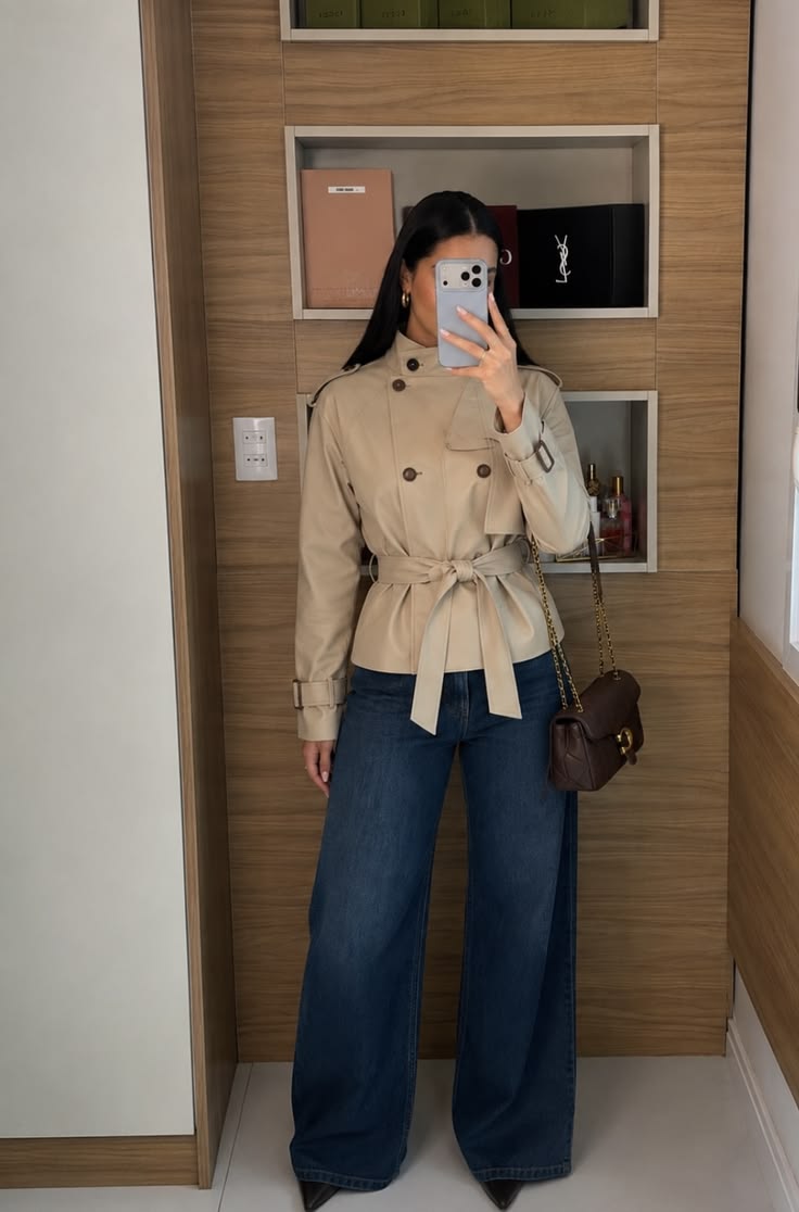 Mulher de cabelos escuros, com um casaco bege estilo trench coat amarrado na cintura, cal&ccedil;a jeans larga e bolsa marrom de al&ccedil;a dourada, tira uma selfie no espelho. Ao fundo, uma parede de madeira com nichos decorativos