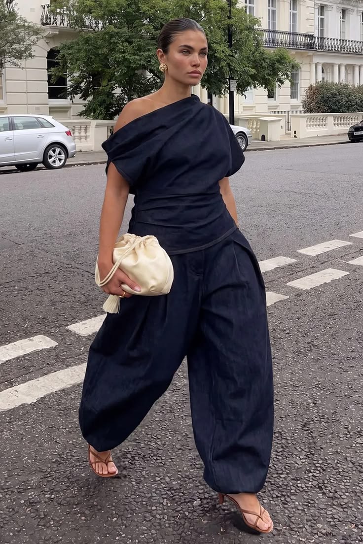Mulher com cabelo preso, blusa preta de um ombro s&oacute; e cal&ccedil;a jeans larga, segurando uma bolsa creme, caminhando em uma rua com pr&eacute;dios brancos ao fundo