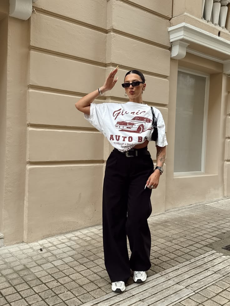 Mulher com &oacute;culos escuros e cabelo preso, vestindo camiseta branca com estampa de carro e cal&ccedil;a preta larga, posa na cal&ccedil;ada com a m&atilde;o na testa