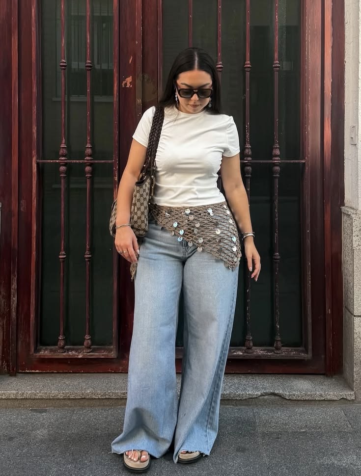 Mulher de cabelos escuros e &oacute;culos de sol, vestindo camiseta branca, cal&ccedil;a jeans larga e cinto de lantejoulas, posa em frente a uma porta de madeira escura com grades