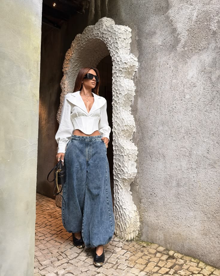Mulher de cabelos castanhos e &oacute;culos escuros, vestindo blusa branca estilo corset e cal&ccedil;a jeans larga, posa em um arco de parede texturizada