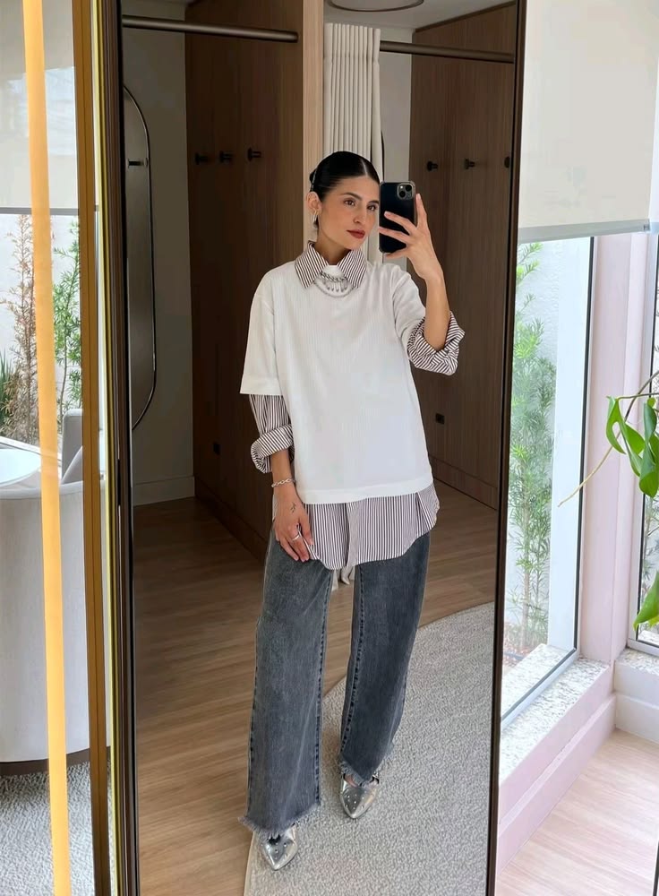 Mulher jovem, de pele clara e cabelos escuros presos, tira uma selfie no espelho. Ela veste uma camiseta branca sobre uma camisa listrada de manga comprida, cal&ccedil;a jeans escura e sapatos prateados. Ao fundo, um quarto com arm&aacute;rios de madeira e uma janela com plantas