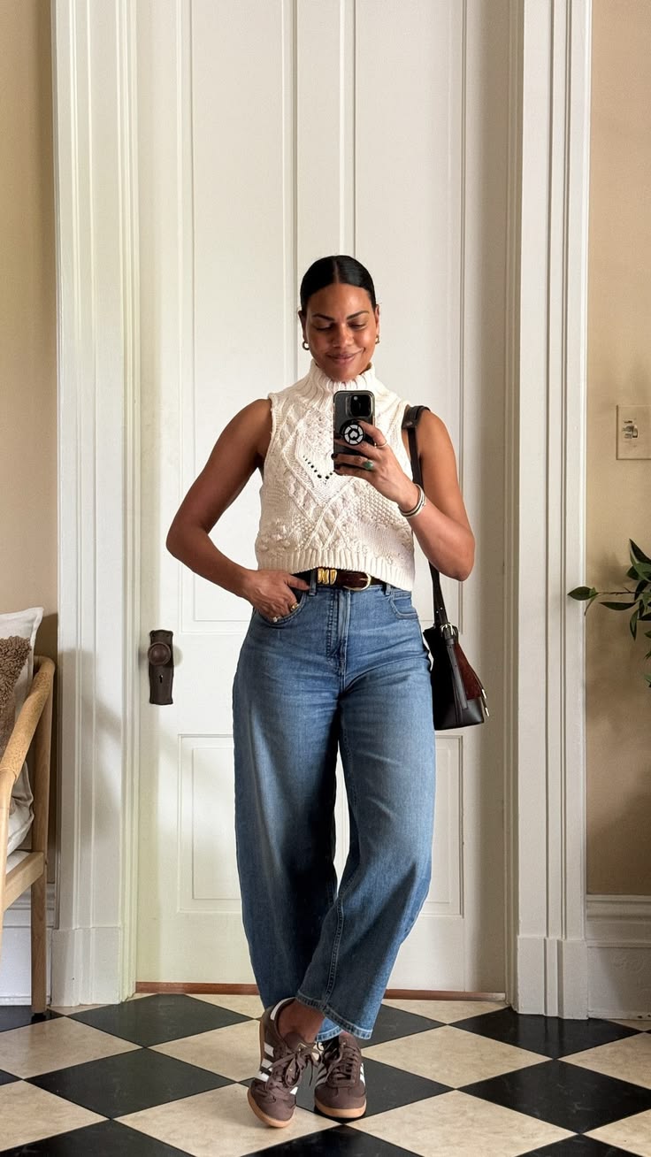 Mulher sorridente, pele morena e cabelo preso, tira selfie no espelho. Veste blusa de tric&ocirc; creme sem mangas, cal&ccedil;a jeans larga, cinto marrom e t&ecirc;nis marrons com listras brancas. Uma bolsa escura est&aacute; pendurada no ombro. O fundo mostra uma porta branca e piso xadrez preto e branco