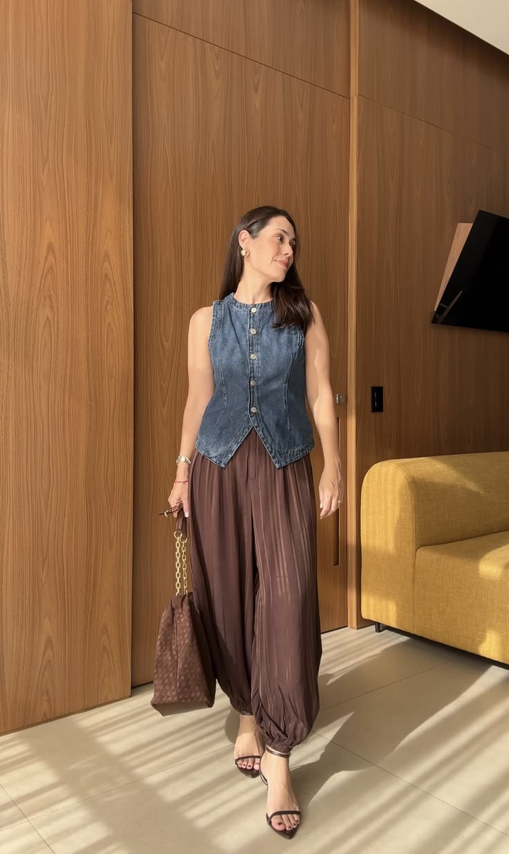Mulher sorridente, de cabelos castanhos soltos, vestindo colete jeans e cal&ccedil;a pantalona marrom, segurando uma bolsa marrom com al&ccedil;a de corrente dourada. Ela est&aacute; em um ambiente com paredes de madeira e um sof&aacute; amarelo ao fundo, com luz natural projetando sombras no ch&atilde;o.