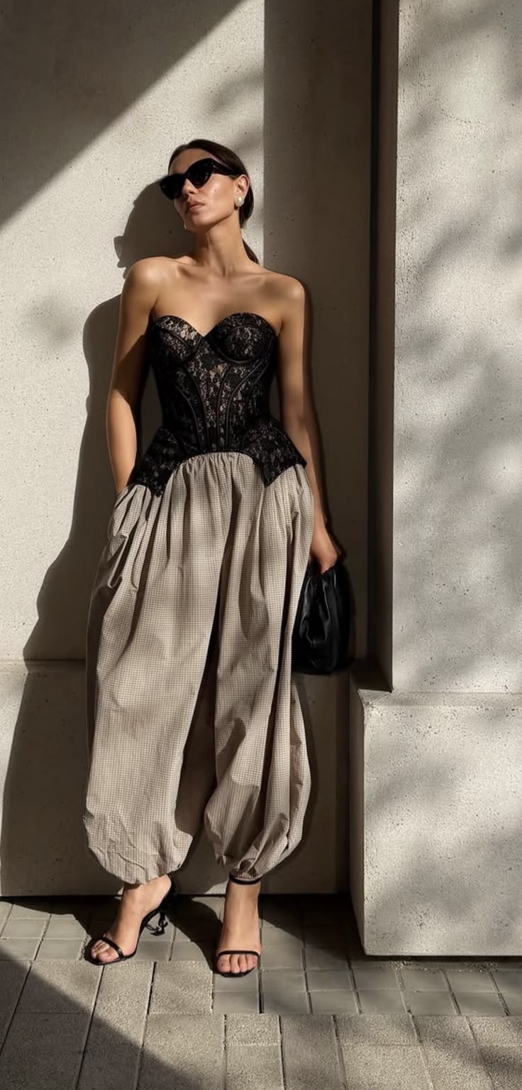 Mulher com &oacute;culos escuros, top corset de renda preta e cal&ccedil;a bege xadrez com volume, posando contra uma parede clara com sombras diagonais. Ela segura uma bolsa preta e usa sand&aacute;lias de salto