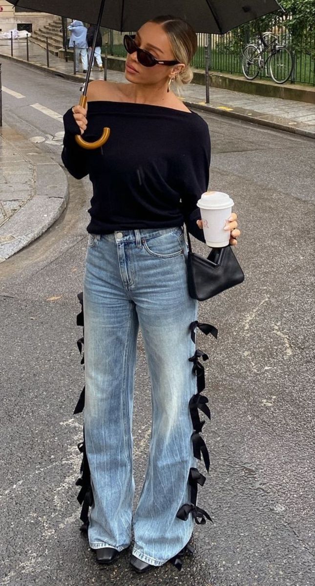 Mulher loira de &oacute;culos escuros e coque, segurando um guarda-chuva preto e um copo de caf&eacute;, vestindo blusa preta ombro a ombro e cal&ccedil;a jeans larga com la&ccedil;os pretos nas laterais, em uma rua molhada