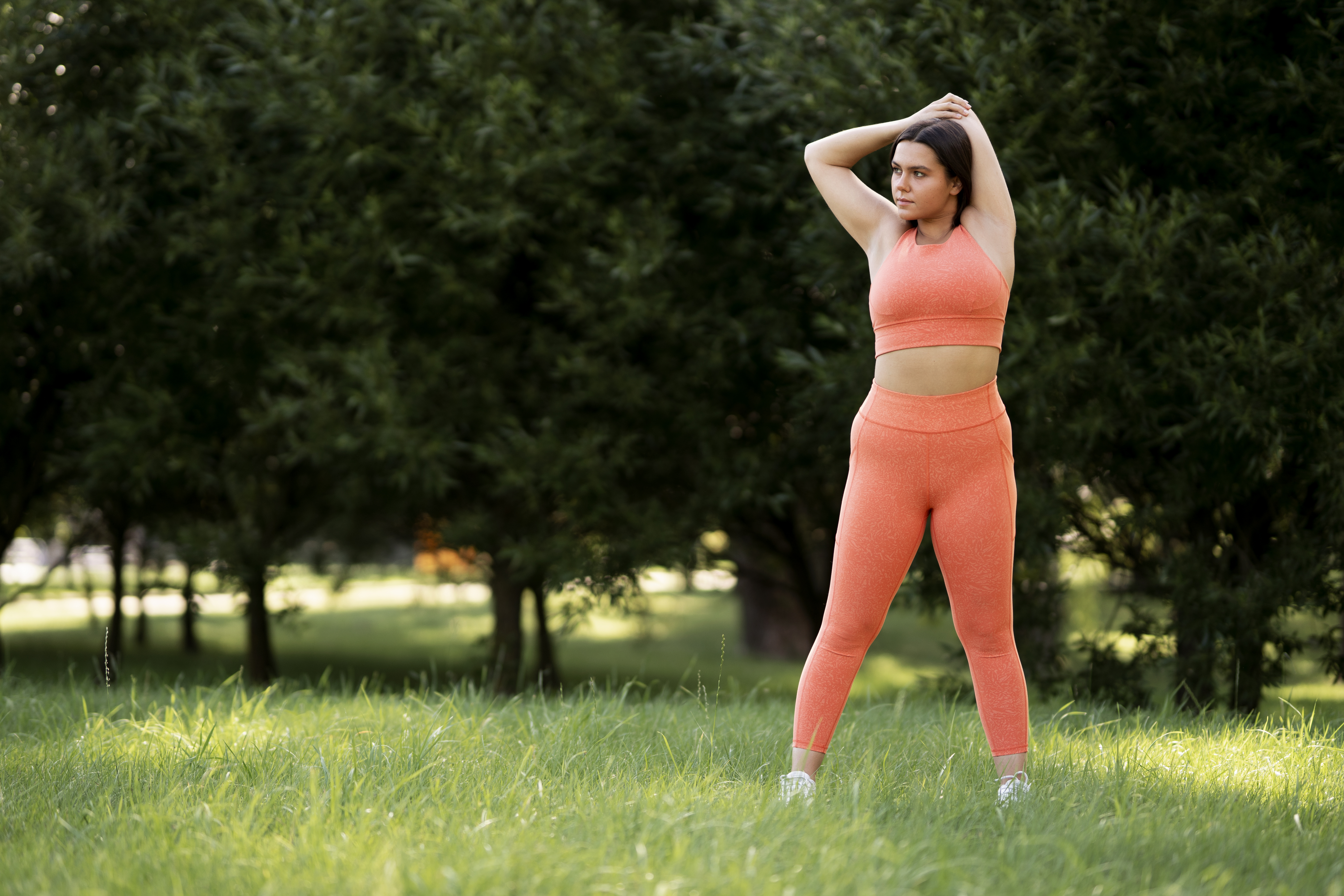 Mulher jovem, de pele clara e cabelo escuro preso, alongando os bra&ccedil;os acima da cabe&ccedil;a, vestindo top e cal&ccedil;a legging laranja, em um gramado verde com &aacute;rvores ao fundo