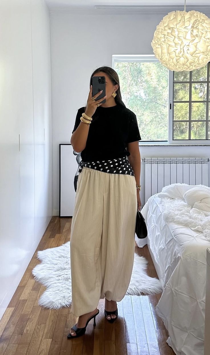 Mulher de cabelo castanho escuro tirando selfie no espelho, vestindo camiseta preta, cal&ccedil;a pantalona bege, len&ccedil;o preto de bolinhas brancas na cintura, sand&aacute;lias pretas de salto, brincos e pulseiras douradas. Ao fundo, quarto com guarda-roupa branco, janela com vista para &aacute;rvores, radiador, cama com edredom branco e tapete de pele sint&eacute;tica branca no ch&atilde;o de madeira