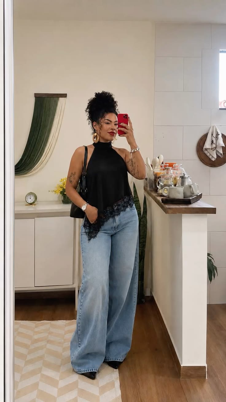 Mulher de pele morena, cabelo crespo preso no alto, blusa preta de gola alta e renda na barra, cal&ccedil;a jeans larga, bolsa preta e brincos dourados, tirando selfie no espelho. Ela est&aacute; em uma cozinha com balc&atilde;o de madeira e azulejos brancos, com objetos de decora&ccedil;&atilde;o e plantas ao fundo