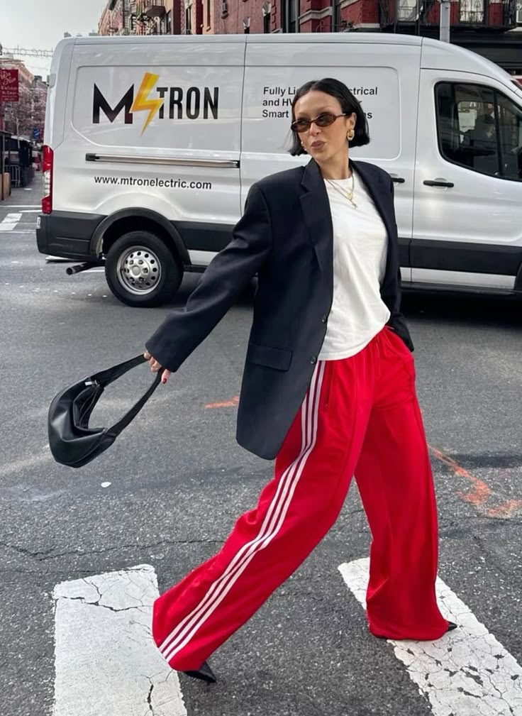 Mulher de cabelo curto e &oacute;culos escuros, vestindo blazer preto, camiseta branca e cal&ccedil;a esportiva vermelha com listras brancas, atravessa a rua segurando uma bolsa preta. Ao fundo, uma van branca da Mtron Electric