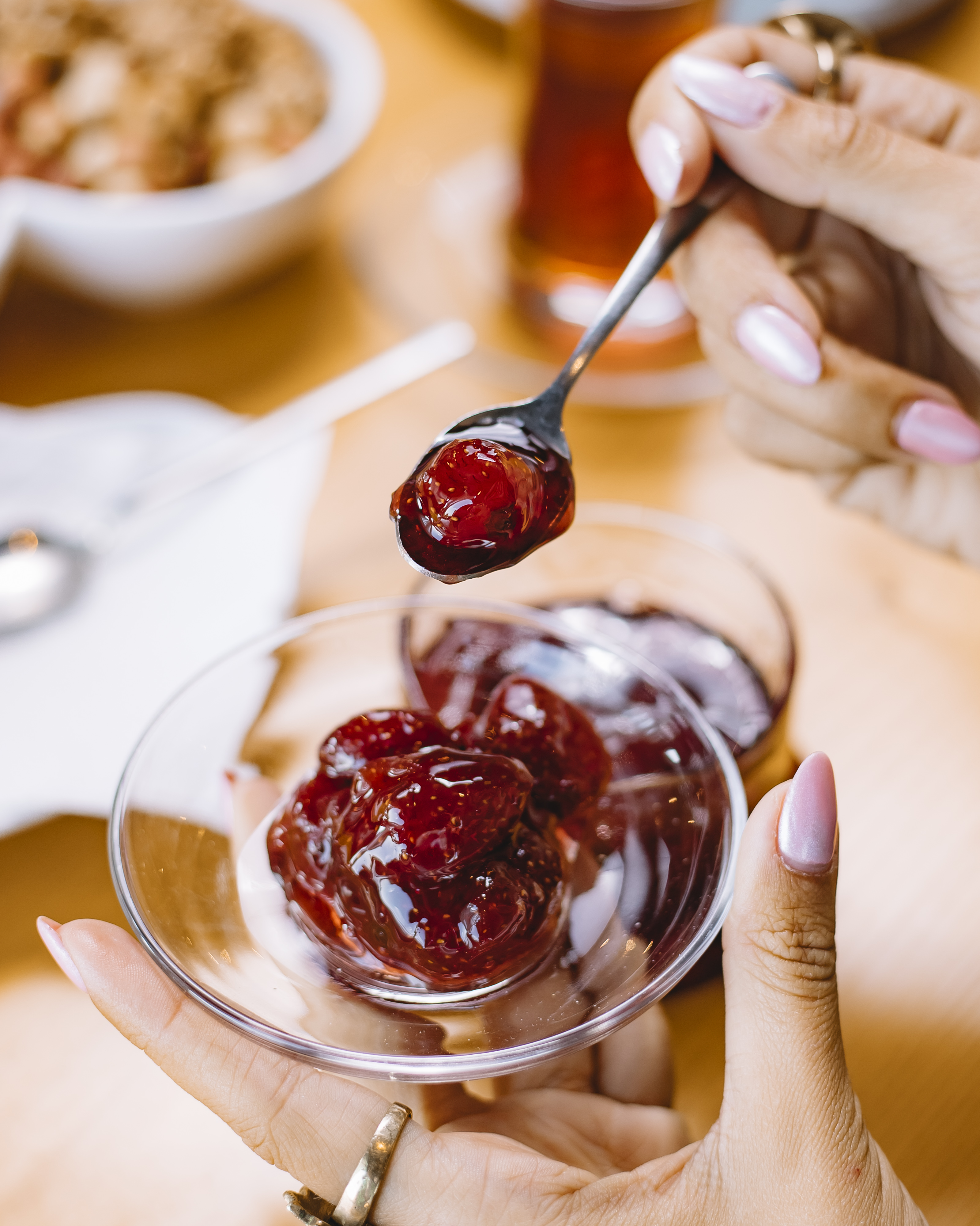 Duas m&atilde;os femininas com unhas longas e rosadas seguram uma tigela de vidro com geleia de morango, enquanto uma colher cheia de geleia &eacute; erguida acima