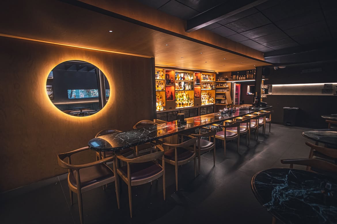 Interior de um bar com ilumina&ccedil;&atilde;o ambiente quente. &Agrave; esquerda, um espelho circular com luz laranja ao redor. Ao centro, um balc&atilde;o comprido de m&aacute;rmore escuro com cadeiras de madeira e estofamento vinho. Ao fundo, prateleiras iluminadas exibem garrafas de bebidas. O teto &eacute; escuro e o ch&atilde;o cinza.