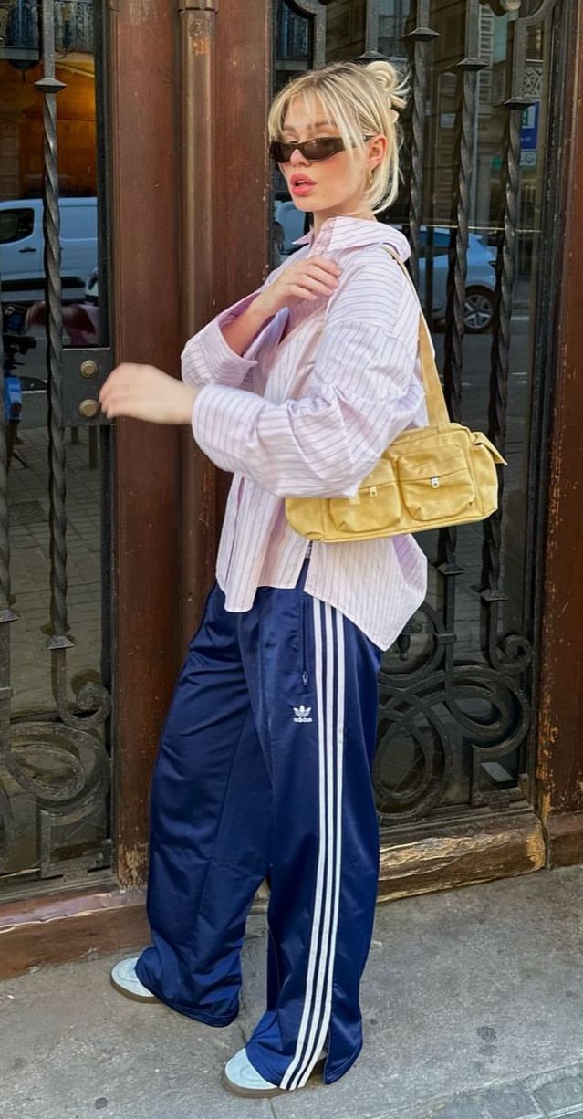 Mulher loira com coque e franja, usando &oacute;culos escuros, camisa listrada rosa e branca, cal&ccedil;a esportiva azul-marinho com listras brancas e bolsa amarela, posando em frente a um port&atilde;o de ferro trabalhado