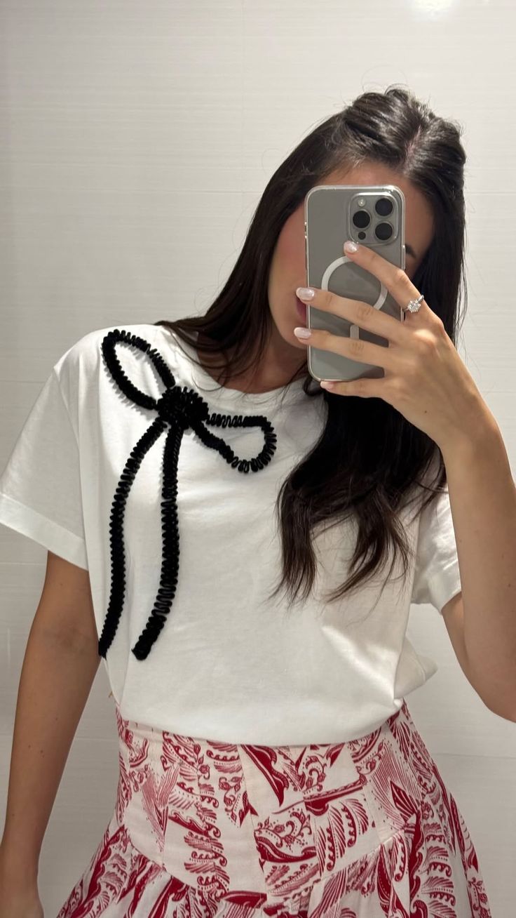 Mulher de cabelos escuros tirando uma selfie no espelho, usando camiseta branca com la&ccedil;o preto texturizado e saia estampada em tons de vermelho e branco. Ela exibe um anel com pedra brilhante no dedo anelar direito e unhas claras