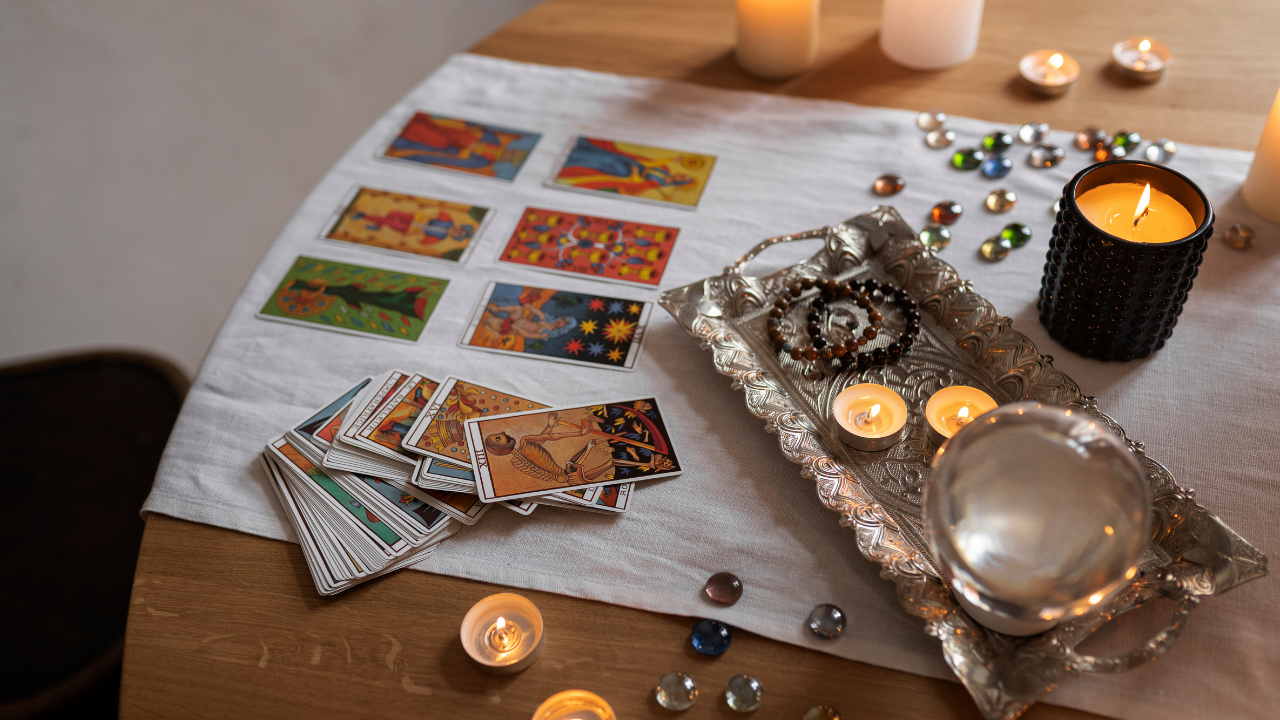 Mesa de madeira com toalha branca, cartas de tarô espalhadas, algumas viradas para cima, e um tabuleiro de prata com velas acesas, pulseiras e uma esfera de cristal, criando uma atmosfera mística