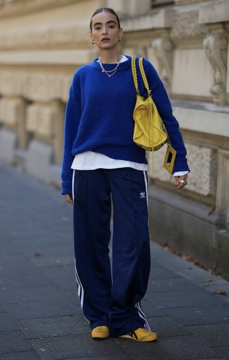 Mulher com cabelo preso, su&eacute;ter azul, camiseta branca, cal&ccedil;a esportiva azul com listras brancas e t&ecirc;nis amarelos, segurando uma bolsa amarela, posando em uma cal&ccedil;ada com um pr&eacute;dio claro ao fundo