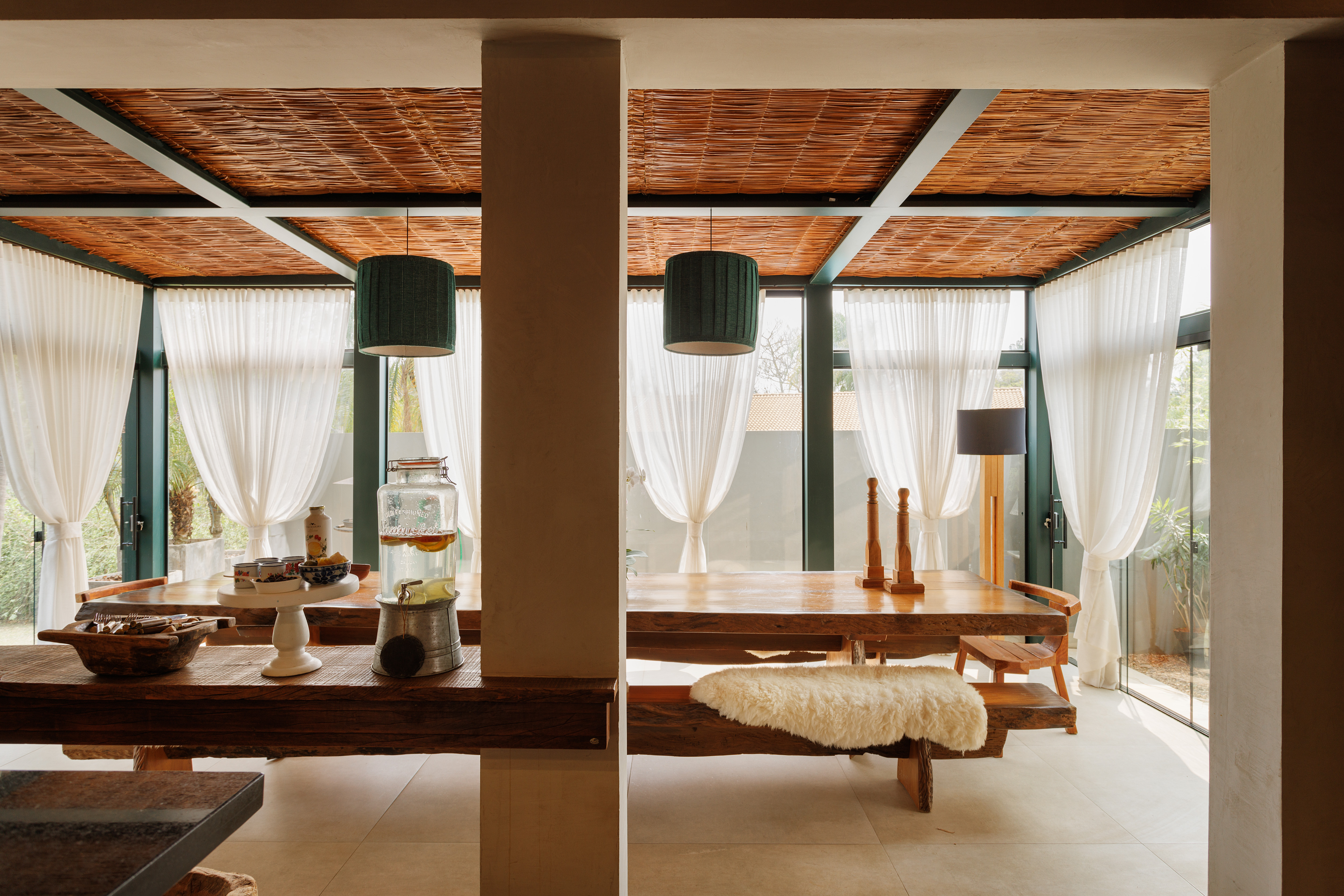 Ambiente interno com duas mesas de madeira r&uacute;stica, bancos e cadeiras. O teto tem acabamento em palha e h&aacute; cortinas brancas transl&uacute;cidas nas grandes janelas. Um pilar central divide o espa&ccedil;o, com lumin&aacute;rias pendentes verdes e um abajur preto. Na mesa da esquerda, um pote de vidro com bebida e tigelas com alimentos. No banco da direita, uma pele de carneiro branca. O piso &eacute; de azulejos claros.