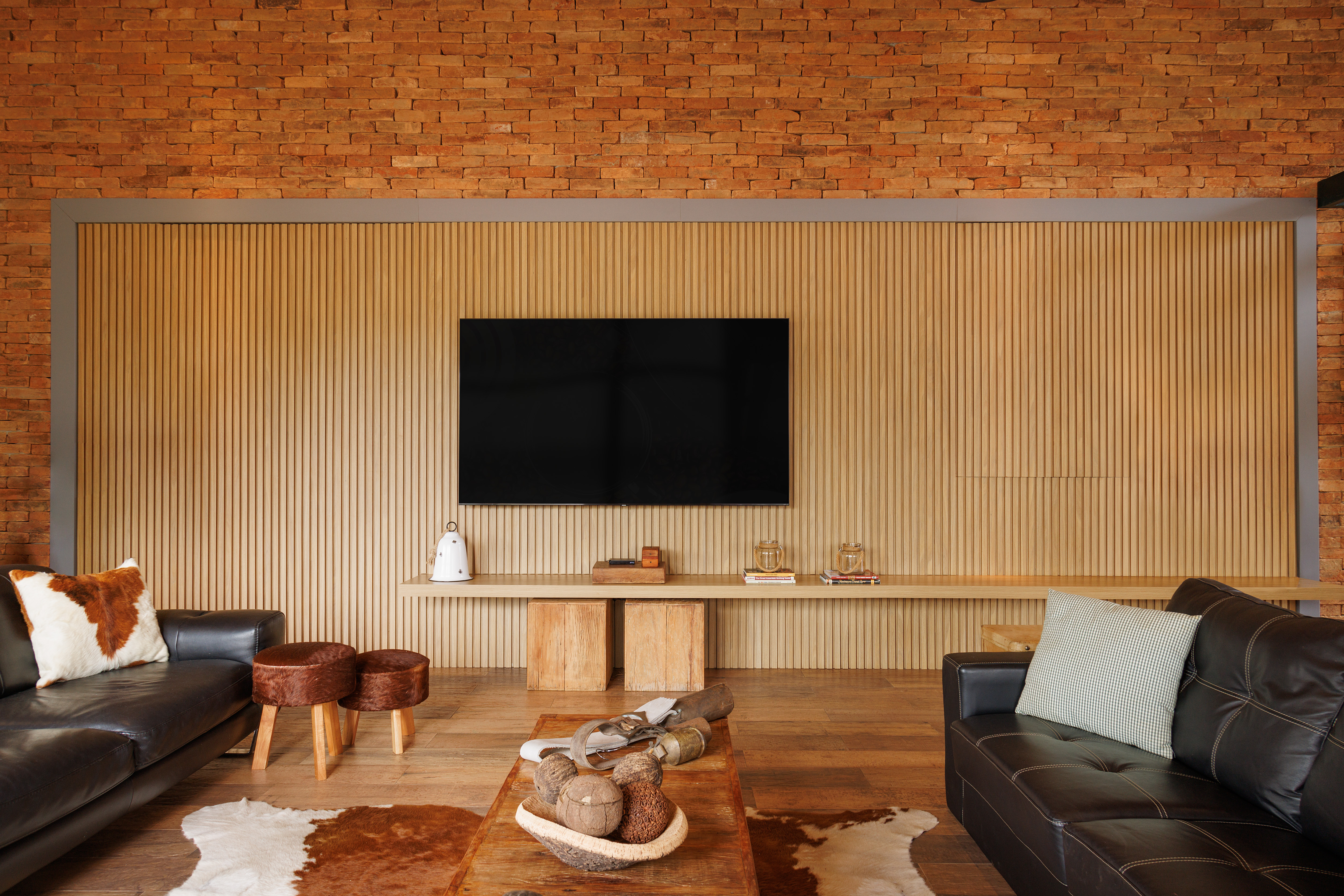 Sala de estar com parede de tijolos aparentes e painel ripado de madeira clara, com uma televis&atilde;o preta centralizada. Abaixo, uma prateleira de madeira com objetos decorativos e dois bancos de madeira r&uacute;stica. Dois sof&aacute;s de couro escuro, um &agrave; esquerda com almofada de pelo, e outro &agrave; direita com almofada xadrez. No centro, uma mesa de centro de madeira com tigela de cocos e tapetes de pele de vaca no ch&atilde;o de madeira