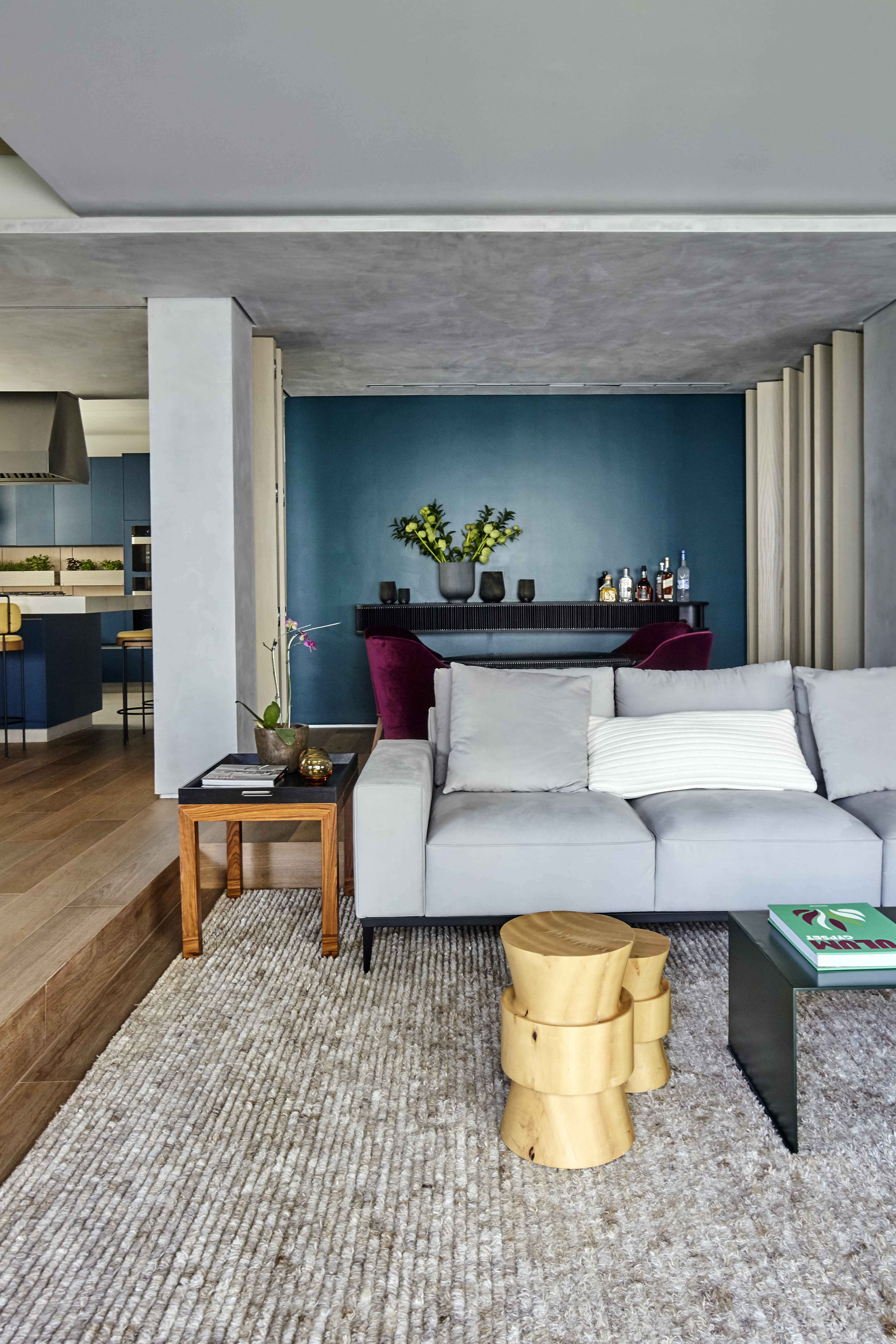 Sala de estar moderna com sof&aacute; cinza, almofadas claras, mesa de centro escura e dois bancos de madeira clara sobre um tapete felpudo. Ao fundo, parede azul-petr&oacute;leo com aparador preto, garrafas e vasos de plantas. &Agrave; esquerda, cozinha azul com bancada e banquetas. O piso &eacute; de madeira e o teto de concreto aparente