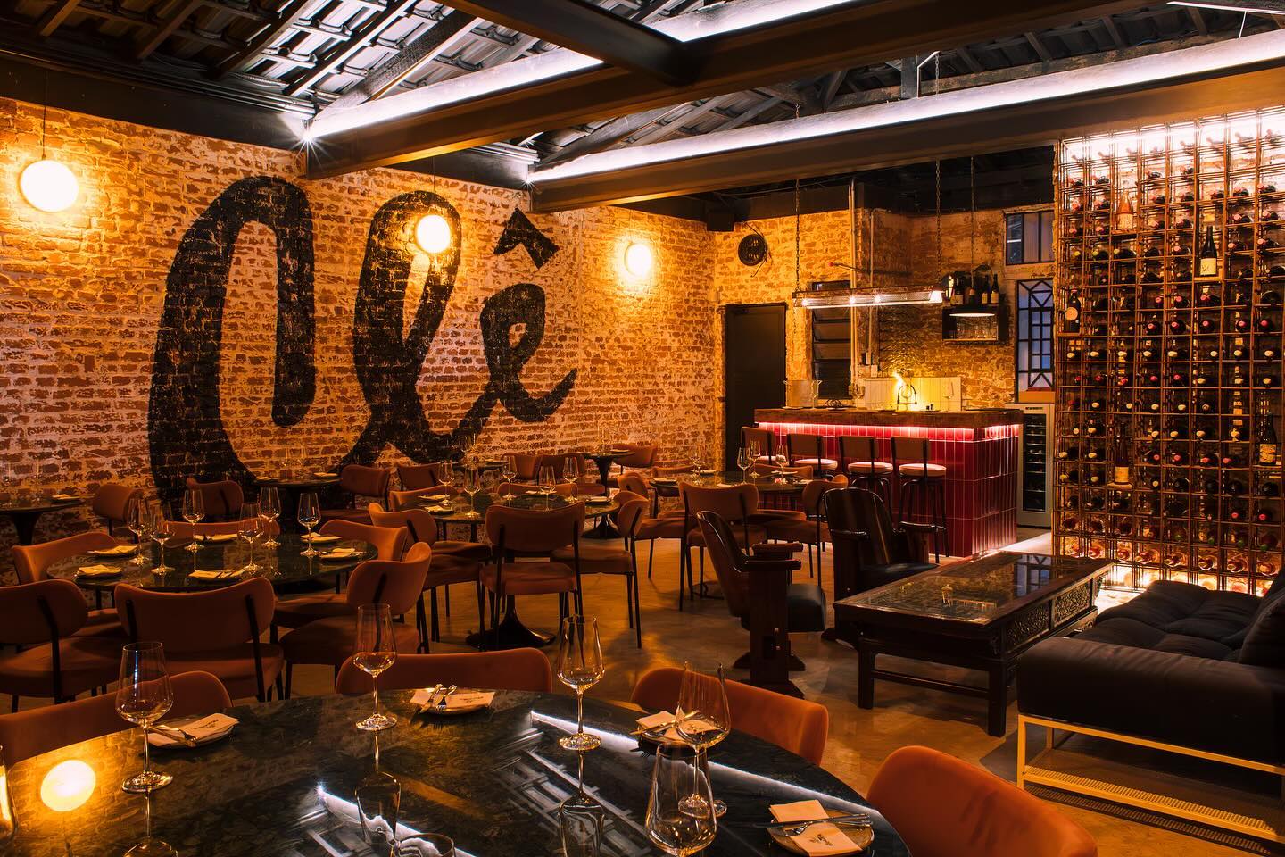 Interior de um restaurante com paredes de tijolos aparentes e a palavra Ol&ecirc; pintada em preto. Mesas redondas com cadeiras de veludo laranja est&atilde;o dispostas pelo sal&atilde;o, com ta&ccedil;as de vinho e pratos. Ao fundo, um bar com balc&atilde;o iluminado em vermelho e uma grande adega de vinhos em grade met&aacute;lica. O ambiente &eacute; acolhedor, com ilumina&ccedil;&atilde;o quente e r&uacute;stica