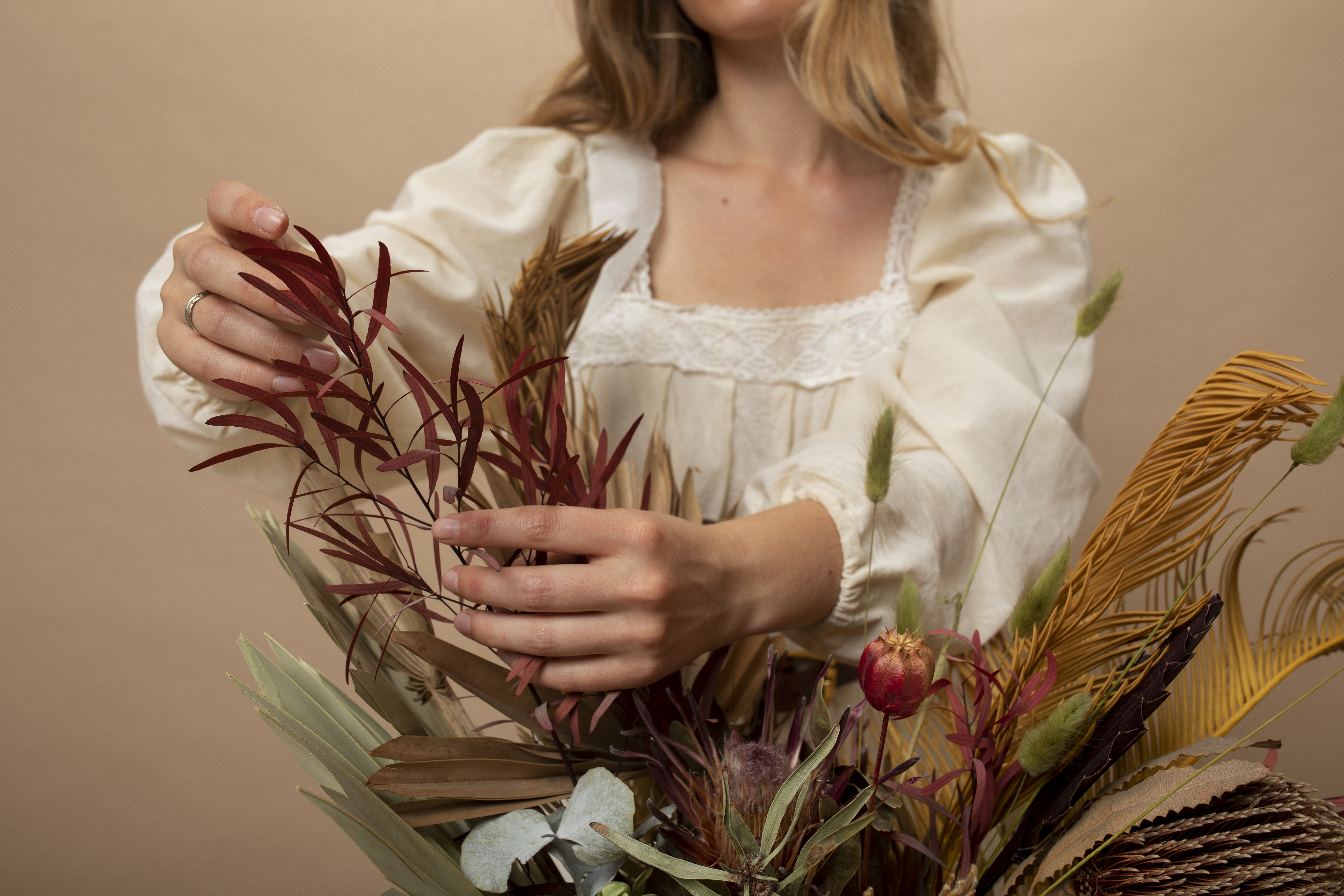 Mulher de blusa creme com renda no decote e mangas bufantes, arrumando um buqu&ecirc; de flores secas e folhagens em tons de vermelho, marrom e verde, com fundo bege