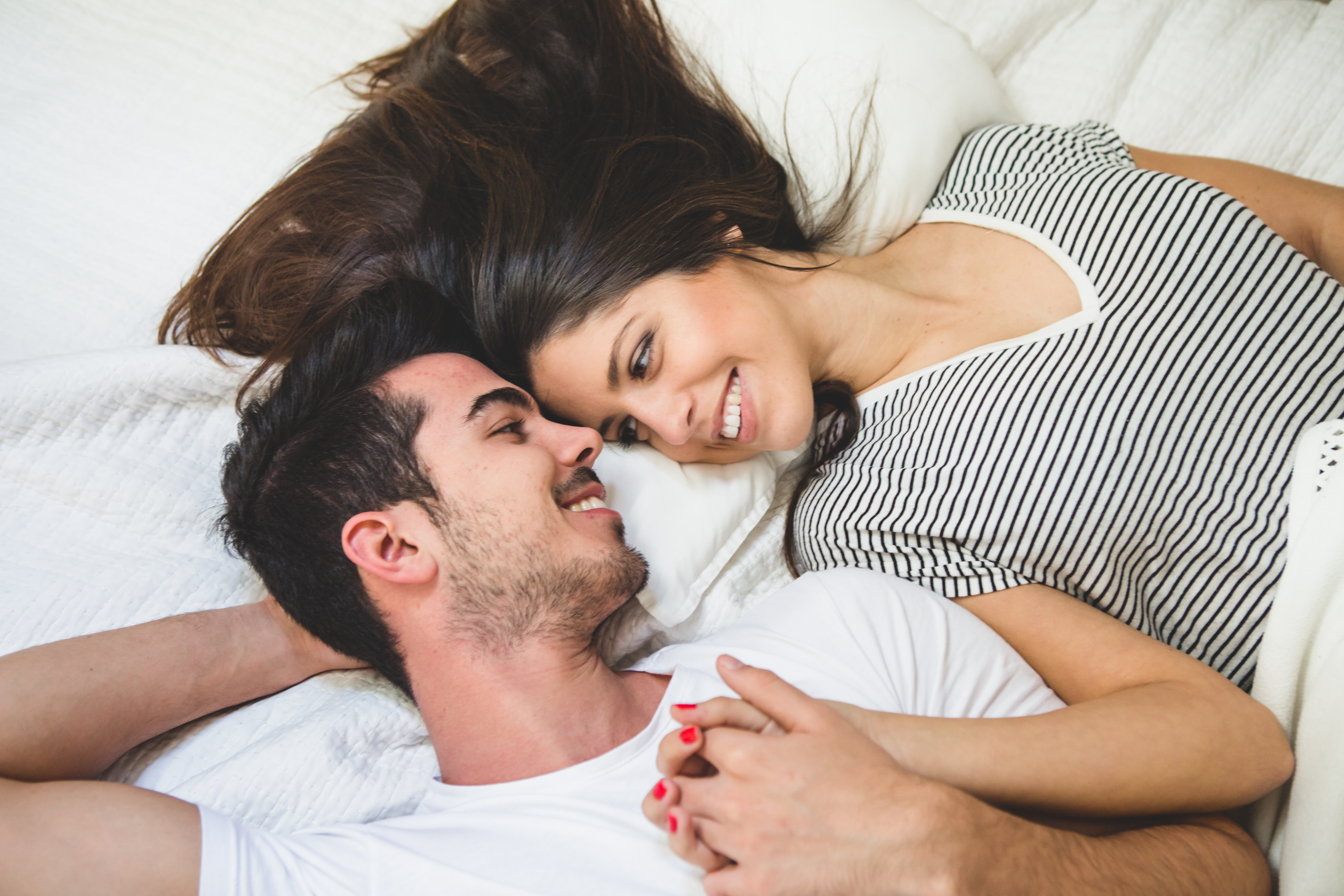Um casal jovem deitado em uma cama branca, sorrindo e se olhando. A mulher, com cabelos longos e escuros, usa uma blusa listrada preta e branca. O homem, com barba rala, veste uma camiseta branca e segura a m&atilde;o dela, que tem unhas vermelhas