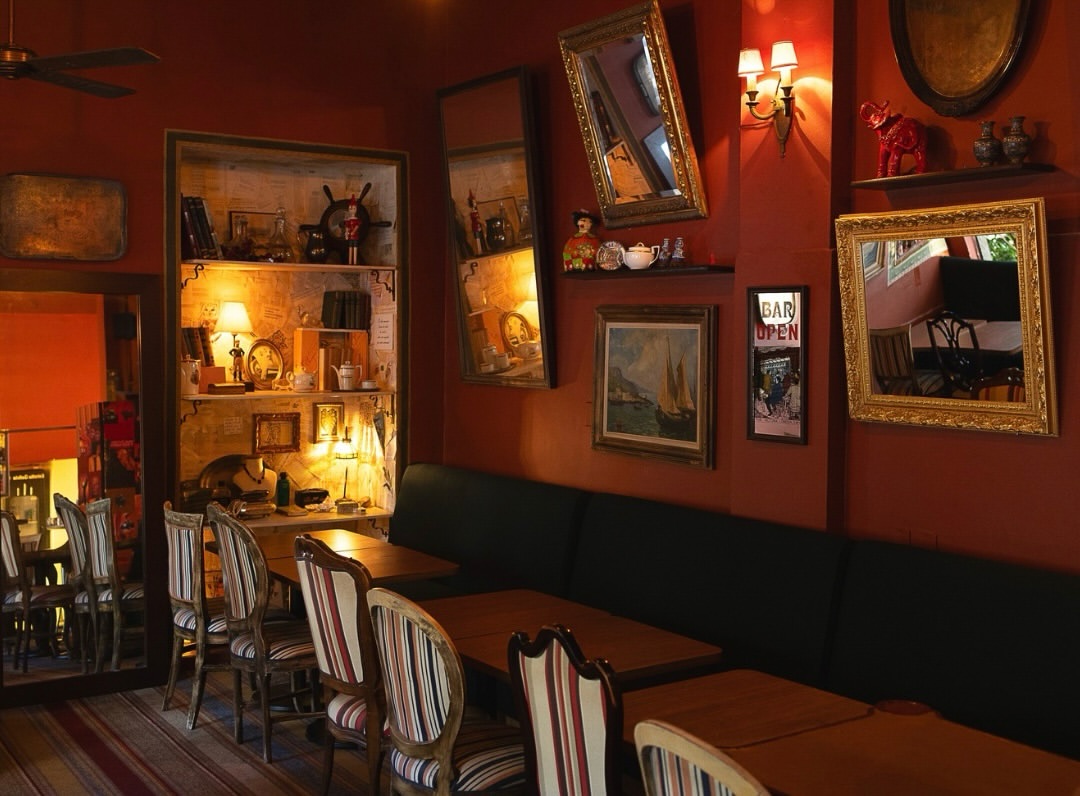 Interior de um bar ou restaurante com paredes vermelhas escuras, mesas de madeira e cadeiras com estofamento listrado. H&aacute; uma estante iluminada com objetos decorativos, espelhos e quadros nas paredes, e um sof&aacute; preto ao longo da parede direita