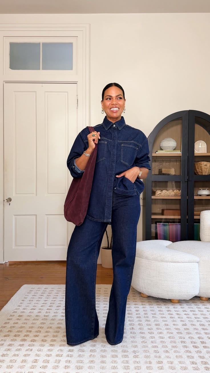 Mulher negra sorridente, cabelo preso, vestindo camisa e cal&ccedil;a jeans escuras, com uma bolsa vinho no ombro, em uma sala com porta branca e arm&aacute;rio preto