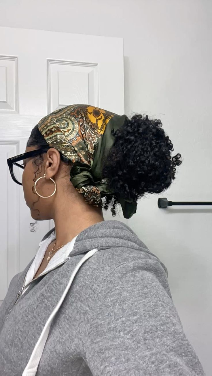Pessoa de pele escura, de perfil, usando &oacute;culos pretos, brincos de argola dourados e um len&ccedil;o estampado em tons de verde e amarelo que prende o cabelo crespo em um coque alto. Veste um moletom cinza com z&iacute;per e capuz, e um colar dourado. O fundo &eacute; uma porta branca e uma parede clara com um suporte preto.