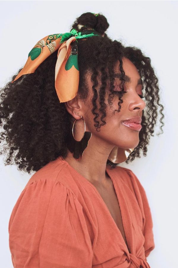Mulher negra de perfil, olhos fechados, cabelo crespo preso com len&ccedil;o laranja e verde, brincos grandes dourados, blusa laranja com decote em V e mangas bufantes